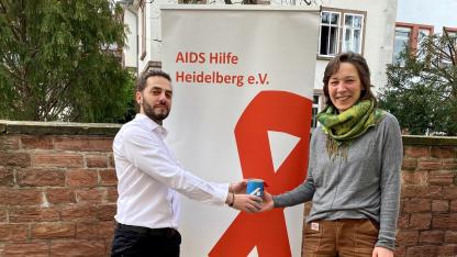 Cosimo Chiarello übergibt an Jennifer Adler eine Spendendose mit einem blauen Etikett. Sie stehen im Freien vor einem Roll-up der AIDS-Hilfe Heidelberg.