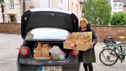 Elke Adler hält eine große Box mit vielen Papiertüten. Auf der Box sind verschiedene frauenspezifische Flyer geklebt. Sie steht neben einem schwarzen Auto. Der Kofferraum ist offen und es sind weitere Boxen mit den gleichen Tüten zu sehen.