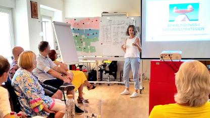 Melanie Ludwig steht vor einer Leinwand und einem Whiteboard und referiert. Hauptamtliche Mitarbeiter*innen und Vorstände der AIDS-Hilfe sitzen um sie herum und hören zu.