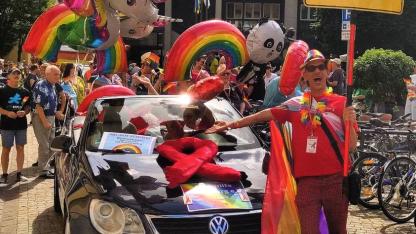 Das AIDS-Hilfe Cabrio in schwarz, geschmückt mit einer roten Schleife. Davor, daneben und dahinter viele Menschen, die bunt gekleidet sind oder unsere roten AIDS-Hilfe Heidelberg T-Shirts tragen. Eine Person trägt das Schild (optisch angelehnt an ein gelbes Ortseingangschild), auf dem AIDS-Hilfe Heidelberg steht. Es sind viele Regenbogen- / Pride Flaggen zu sehen