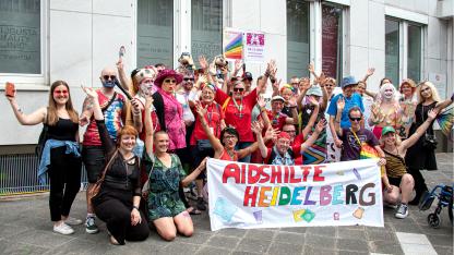 Gruppenfoto: etwa 30 Menschen stehen zusammen und heben die Arme. Sie sind zum Teil geschminkt und bunt angezogen. Einige tragen die roten Aidshilfe-T-Shirts. Die vorderen Menschen knien auf dem Boden. Sie halten ein Banner, auf dem "AIDSHILFE HEIDELBERG" steht.