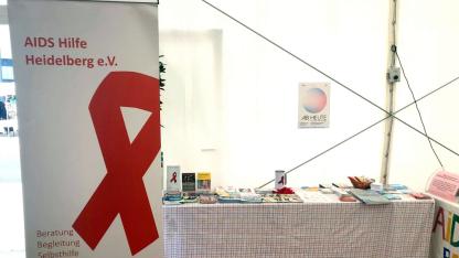 Es ist ein Infostand mit diversem Infomaterial zu sehen. Davor steht das Roll-up der AIDS-Hilfe Heidelberg.