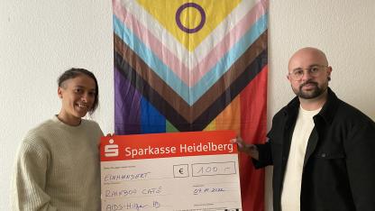 Eva (Rainbowcafe) übergibt Noah einen Spendenscheck. Die beiden stehen vor einer weißen Wand, an der eine Progress Flagge hängt.
