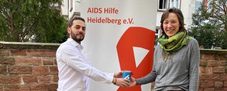 Cosimo Chiarello übergibt an Jennifer Adler eine Spendendose mit einem blauen Etikett. Sie stehen im Freien vor einem Roll-up der AIDS-Hilfe Heidelberg.