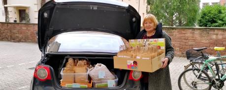 Elke Adler hält eine große Box mit vielen Papiertüten. Auf der Box sind verschiedene frauenspezifische Flyer geklebt. Sie steht neben einem schwarzen Auto. Der Kofferraum ist offen und es sind weitere Boxen mit den gleichen Tüten zu sehen.