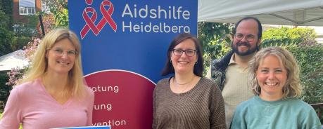 Gruppenfoto: Carolin Gast, Juliane Schurig, Peter Abelmann und Mahela Stock stehen vor einem Roll-Up der Aidshilfe. Frau Gast von BD hält ein Plakat des Angebots "Checkpoint" hoch. Im Hintergrund blauer Himmel und Häuser.