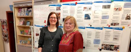 Juliane und Elke stehen vor einem Board mit einem Zeitstrahl zur Geschichte der HIV Epidemie und der Aidshilfe Heidelberg