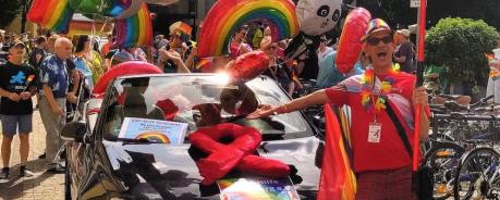 Das AIDS-Hilfe Cabrio in schwarz, geschmückt mit einer roten Schleife. Davor, daneben und dahinter viele Menschen, die bunt gekleidet sind oder unsere roten AIDS-Hilfe Heidelberg T-Shirts tragen. Eine Person trägt das Schild (optisch angelehnt an ein gelbes Ortseingangschild), auf dem AIDS-Hilfe Heidelberg steht. Es sind viele Regenbogen- / Pride Flaggen zu sehen