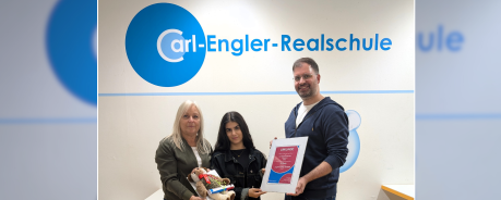 Patrick steht mit anderen Menschen vor einer Wand, an der das Logo der Carl Engler Realschule ist.