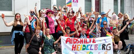 Gruppenfoto: etwa 30 Menschen stehen zusammen und heben die Arme. Sie sind zum Teil geschminkt und bunt angezogen. Einige tragen die roten Aidshilfe-T-Shirts. Die vorderen Menschen knien auf dem Boden. Sie halten ein Banner, auf dem "AIDSHILFE HEIDELBERG" steht.