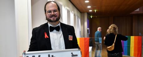 Peter steht vor einem Plakat mit der Aufschrift "Welt-Aids-Tag". Er ist im Theater und tr&auml;gt einen Anzug mit roter Schleife.