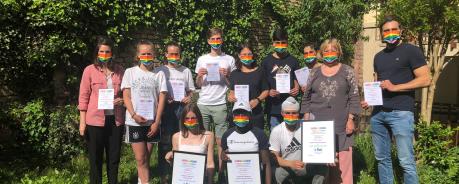 Gruppenbild der Urkundenübergabe der Spendenaktion "Jugend zeigt Schleife": Schüler*innen und Lehrer*innen stehen mit unserer MItarbeiterin Elke Adler zusammen. Alle tragen Regenbogenmasken und halten Urkunden mit verschiedenen Platzierungen.
