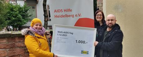 Drei Personen stehen auf dem Bild vor einem Plakat der AIDS Hilfe Heidelberg. Eine Person in einem gelben Mantel übergibt den Mitarbeiter*innen der AIDS-Hilfe einen großen Scheck über 1000€