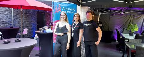 Gruppenbild: Juliane steht mit Chrissi und Daniel von Kinky Escapes vor einem Roll up der Aidshilfe 