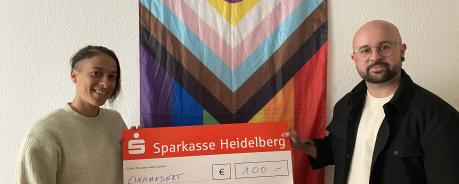 Eva (Rainbowcafe) übergibt Noah einen Spendenscheck. Die beiden stehen vor einer weißen Wand, an der eine Progress Flagge hängt.
