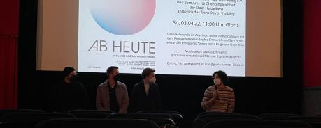 Das Bild zeigt vier Personen, die vor einer Kinoleinwand stehen. Die Personen haben ein Mikrofon und beantworten die Fragen des Publikums. Auf der weißen Leinwand sieht man eine hellblau-rosa farbene Kugel, darunter der Titel des gezeigten Films (Ab heute).