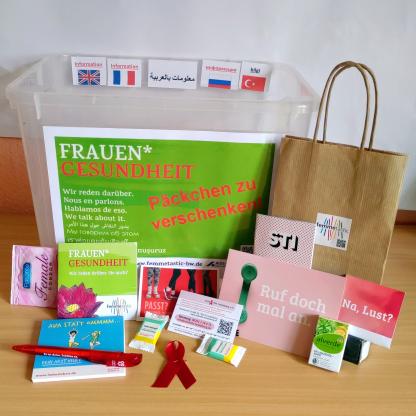 Es liegen viele bunte Flyer und Give-aways rund um Frauengesundheit auf einem Tisch: Ein Femidom, ein Kondom mit dem Slogan: "Na, Lust?", ein Flyer zu STI, eine Postkarte mit dem Slogan: "Ruf doch mal an.", unsere Visitenkarte, Traubenzucker und vieles mehr.