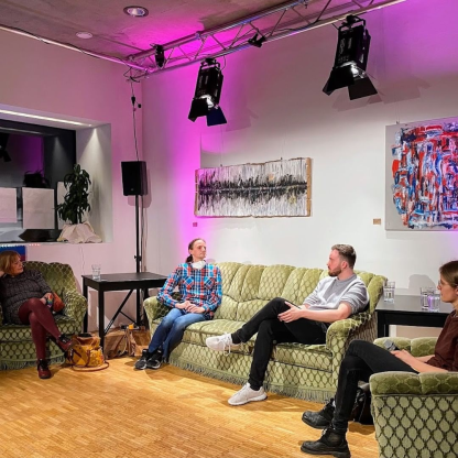 vier Personen sitzen mit Abstand auf drei olivgrünen Sofas. Sie unterhalten sich angeregt, aber entspannt. Sie werden von Scheinwerfern mit pinkem Licht beleuchtet. An der Wand im Hintergrund hängen Kunstwerke.