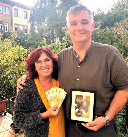 Thomas Grundt steht mit Martina Häusler auf der Terasse der AIDS-Hilfe. Martina hält 1.000 € in der Hand und Thomas ein Bild seines verstorbenen Ehemannes Harald.