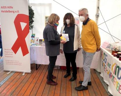 Links im Bild steht das Roll-up der AIDS-Hilfe Heidelberg. Daneben steht eine Gruppe von drei Menschen. Alle tragen Masken und sind ins Gespräch vertieft. Eine davon ist Elke Adler. Sie ist unsere Mitarbeiterin und Mitglied von AIDS und Kinder e.V. Sie hält Broschüren in der Hand. Hinter den Personen ist ein Infostand, auf dem viel Infomaterial liegt, das nicht genau zu erkennen ist.