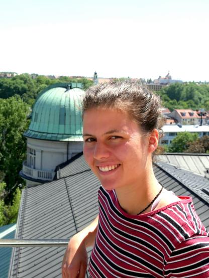 Selfie unserer Praktikant_in Charlie: Charlie hat braune Haare, trägt ein T-Shirt mit rot/weiß/blauen Streifen und lächslt in die Kamera. Charlie steht offenbar auf einem Turm: Im Hintergrund sind Gebäude, Bäume und Himmel zu sehen.