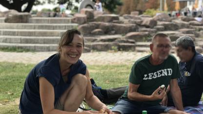 Picknick: Jennifer Adler lächelt in die Kamera. Vor ihr ist eine Picknickdecke mit Essen. Neben ihr sind Menschen, deren Gesichter gepixelt sind.
