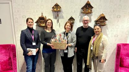  Gruppenbild: Claudia Bach (Reckitt Benckiser), Juliane Schurig (AIDS-Hilfe), Manuela Flühr (Reckitt Benckiser), Peter Spuhler (AIDS-Hilfe), Spasenija Bradic (Reckitt Benckiser). Sie stehen vor einer Wand, die mit Kuckucksuhren geschmückt ist. Links und rechts stehen pinke Sessel. Juliane Schurig und Manuela Flühr halten einen Geschenkkorb.