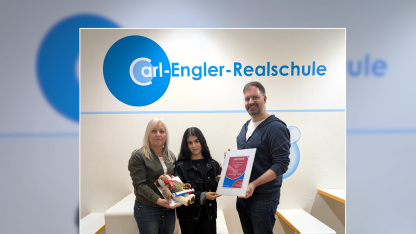 Patrick steht mit anderen Menschen vor einer Wand, an der das Logo der Carl Engler Realschule ist.