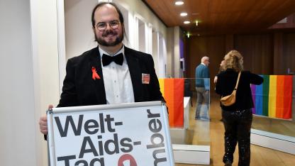 Peter steht vor einem Plakat mit der Aufschrift "Welt-Aids-Tag". Er ist im Theater und tr&auml;gt einen Anzug mit roter Schleife.