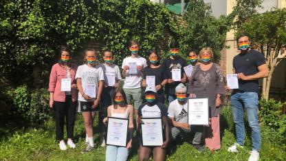Gruppenbild der Urkundenübergabe der Spendenaktion "Jugend zeigt Schleife": Schüler*innen und Lehrer*innen stehen mit unserer MItarbeiterin Elke Adler zusammen. Alle tragen Regenbogenmasken und halten Urkunden mit verschiedenen Platzierungen.