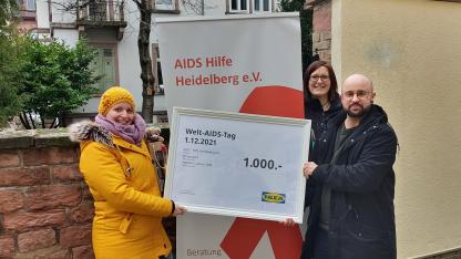 Drei Personen stehen auf dem Bild vor einem Plakat der AIDS Hilfe Heidelberg. Eine Person in einem gelben Mantel übergibt den Mitarbeiter*innen der AIDS-Hilfe einen großen Scheck über 1000€