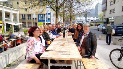 14 Personen sitzen um einen großen Tisch im Freien. Sie sind sehr gut gelaunt und warten auf ihr Essen.