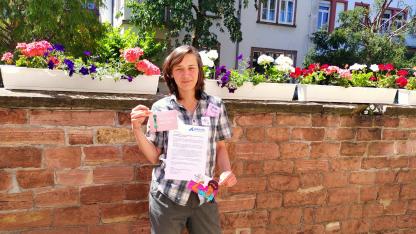 Jennifer Adler steht vor eine Mauer. Sie hält die Pressemitteilung der Aidshilfe Ba-Wü in der Hand und Postkarten der Aktion "Femmetastic".