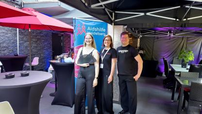 Gruppenbild: Juliane steht mit Chrissi und Daniel von Kinky Escapes vor einem Roll up der Aidshilfe 