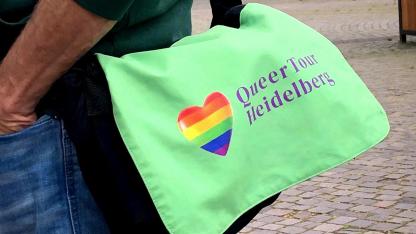 Im Fokus steht eine grüne Umhängetasche. Sie ziert der Schriftzug "QueerTour Heidelberg" und ein Herz in Regenbogenfarben. Sie wird von einer Person in Jeans und dunkelgrünem Pullover getragen. Die Person ist jedoch nur in Ausschnitten zu sehen. Im Hintergrund sind Bäume, Gebäude und Kopfsteinpflaster.
