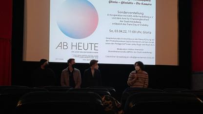Das Bild zeigt vier Personen, die vor einer Kinoleinwand stehen. Die Personen haben ein Mikrofon und beantworten die Fragen des Publikums. Auf der weißen Leinwand sieht man eine hellblau-rosa farbene Kugel, darunter der Titel des gezeigten Films (Ab heute).